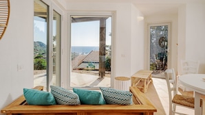 Una habitación con un banco de madera, una mesa y sillas, con vista al mar y un edificio de techo a dos aguas.