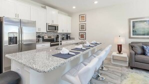 A modern kitchen with white cabinets, stainless steel appliances, and a granite countertop. There are bar stools and a small seating area with a lamp. - Resort acess - Last minute deal (Kissimmee)