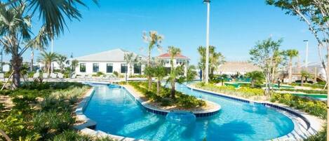A swimming pool with a curved design, surrounded by palm trees and greenery, with buildings in the background.