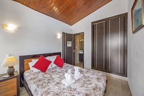 A hotel room with a wooden ceiling, a large bed with floral bedding, red pillows, a nightstand with a lamp, and a wardrobe. - Villa Elo by Villa Plus (Albufeira)