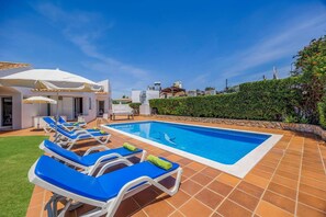 Una piscina con sillas de descanso y sombrillas, rodeada por un patio empedrado y un seto.
