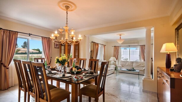 A dining area with a chandelier, wooden chairs, a glass table, and a view of the outdoors. - Villa Rosa by Villa Plus (Olhos de Água)