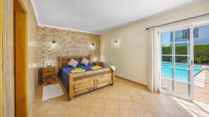 A bedroom with a wooden bed, a nightstand, a wall-mounted lamp, and a view of a swimming pool through a sliding glass door. - Villa Teotel by Villa Plus (Almancil)