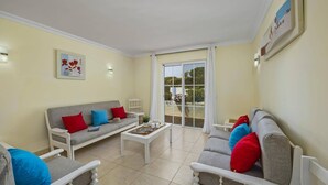 Un salon avec un canapé gris, un fauteuil blanc et une table basse. Une porte-fenêtre coulissante donne accès à un espace extérieur.
