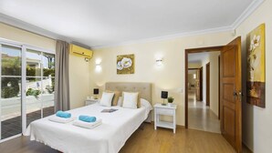Une chambre à coucher avec un grand lit, deux tables de chevet, une commode, un tableau et une vue sur l’extérieur, visible à travers une porte-fenêtre coulissante.