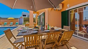 A poolside dining area with a table set for four, surrounded by wooden chairs, under a large umbrella. - Villa Bali by Villa Plus (Albufeira)