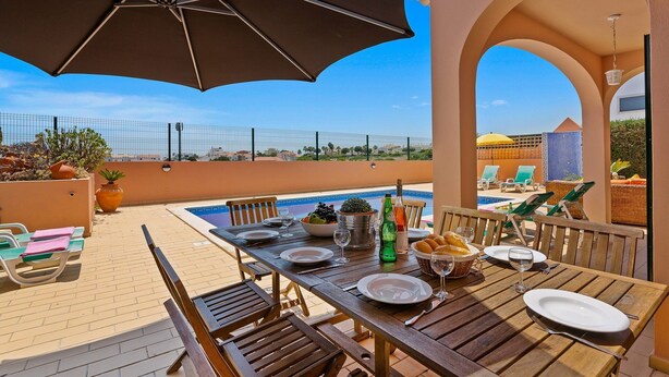 An outdoor dining area with a pool, wooden tables, and chairs, overlooking a clear sky and distant buildings. - Villa Bali by Villa Plus (Albufeira)