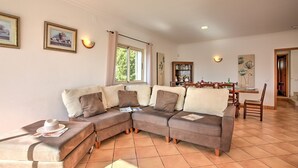 A living room with a sectional sofa, a dining table with chairs, a bookshelf, and a window with curtains. - Villa Papi by Villa Plus (Albufeira)