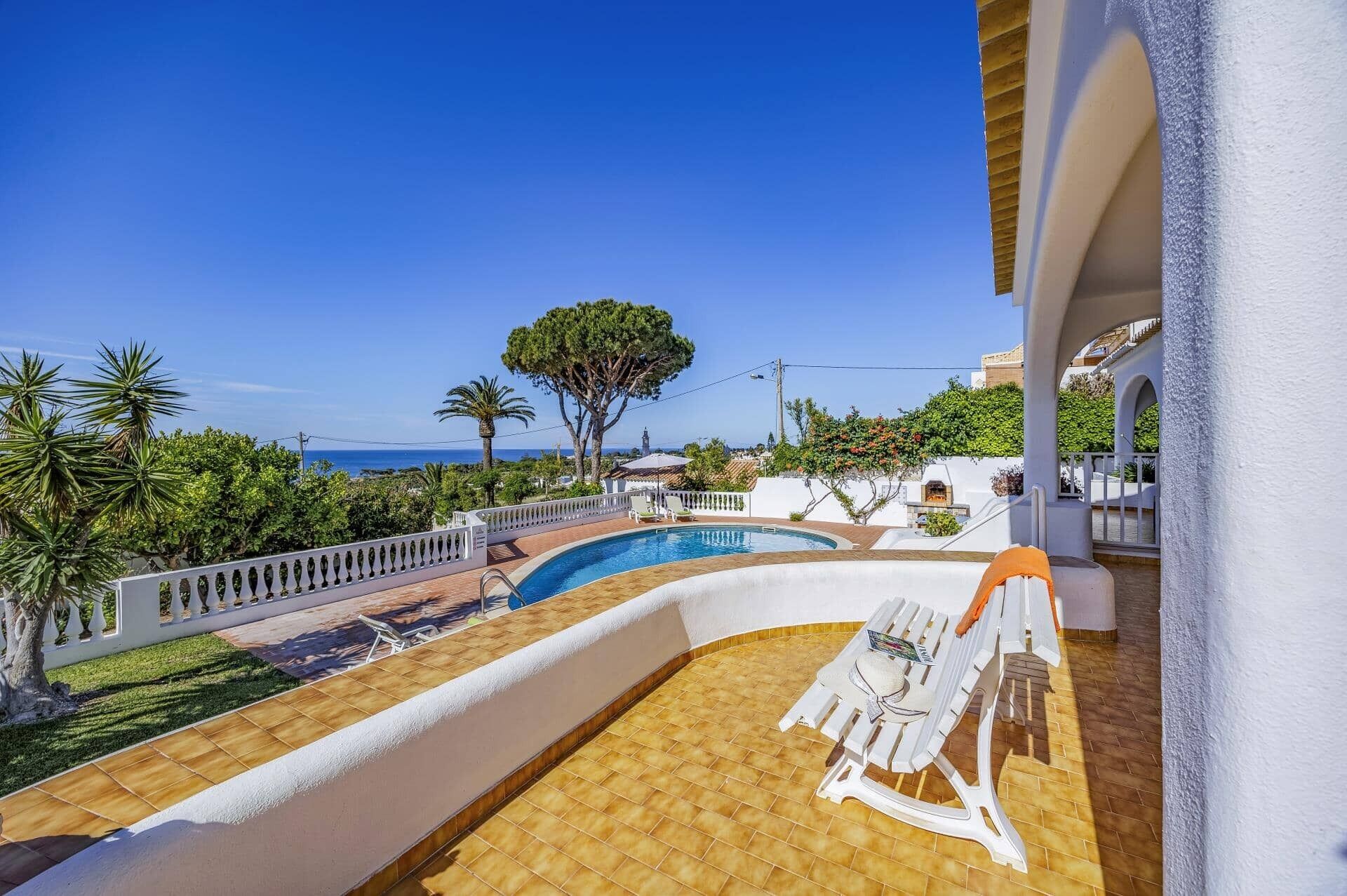 Zona de piscina con vista al mar, un edificio blanco y un árbol.