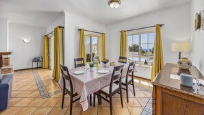 Comedor con una mesa dispuesta para cuatro, sillas, una mesita auxiliar con lámpara y vistas amplias a través de grandes ventanales.