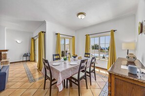 A dining area with a table set for four, chairs, a small side table with a lamp, and a view through large windows. - Villa Primavera by Villa Plus (Albufeira)
