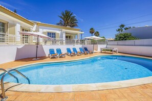 A swimming pool with lounge chairs and umbrellas, surrounded by a white building with a balcony. - Villa Primavera by Villa Plus (Albufeira)