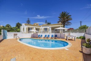 A swimming pool with lounge chairs, a covered seating area, and a building with a balcony. - Villa Primavera by Villa Plus (Albufeira)