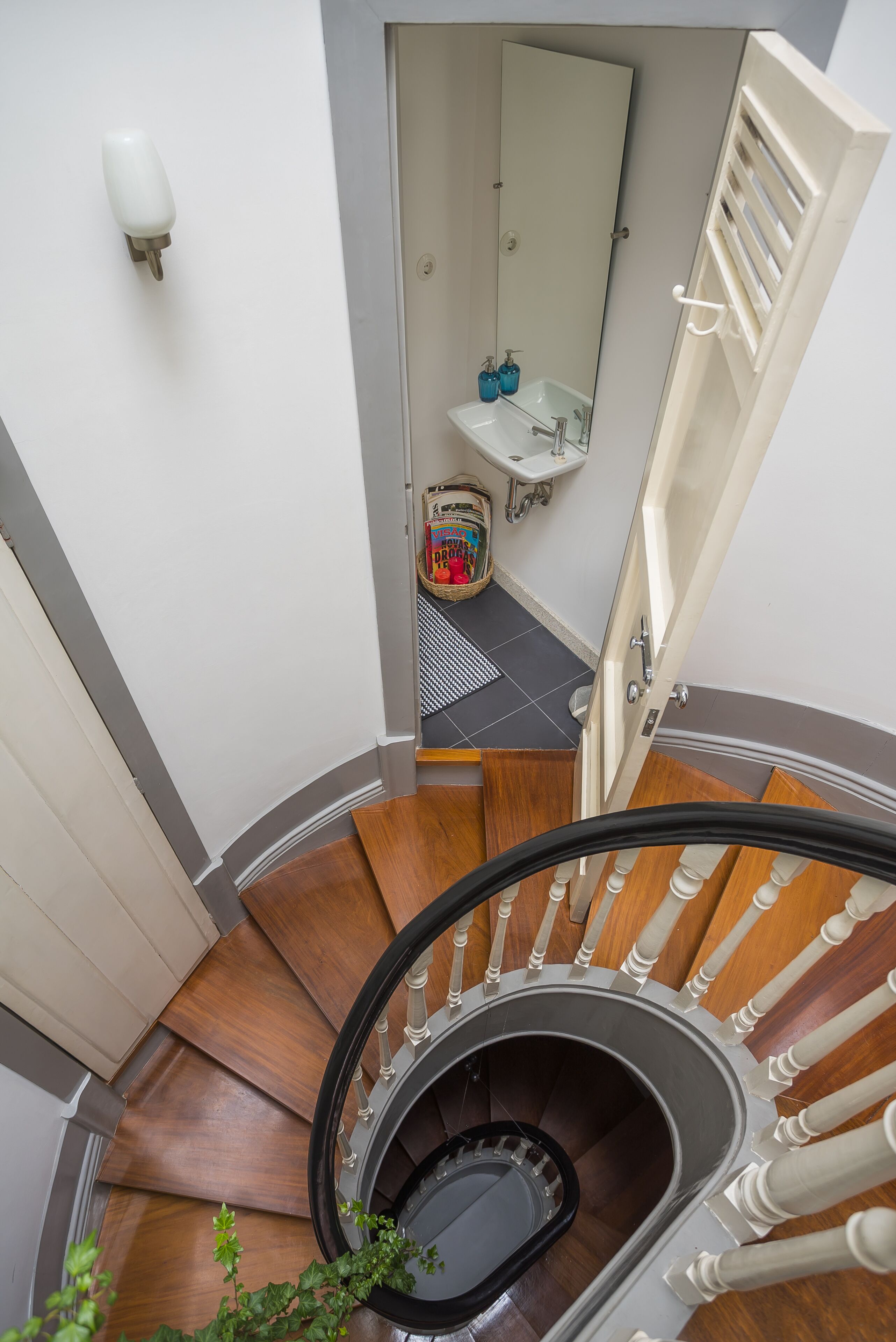 A spiral staircase with wooden steps and white balusters, leading to a bathroom with a sink, mirror, and toiletries.