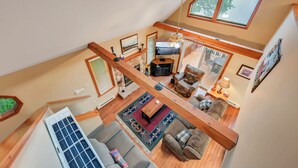 A spacious loft-style living area with a high ceiling, wooden beams, a fireplace, and a large window overlooking a snowy landscape.