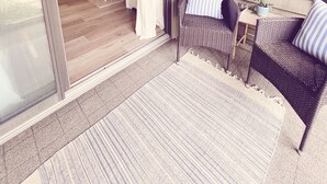 Ein überdachter Patio-Bereich mit Korbmöbeln, einem kleinen Tisch mit einer Pflanze und einer Glastür, die zu einem Innenraum mit Esstisch und Stühlen führt.