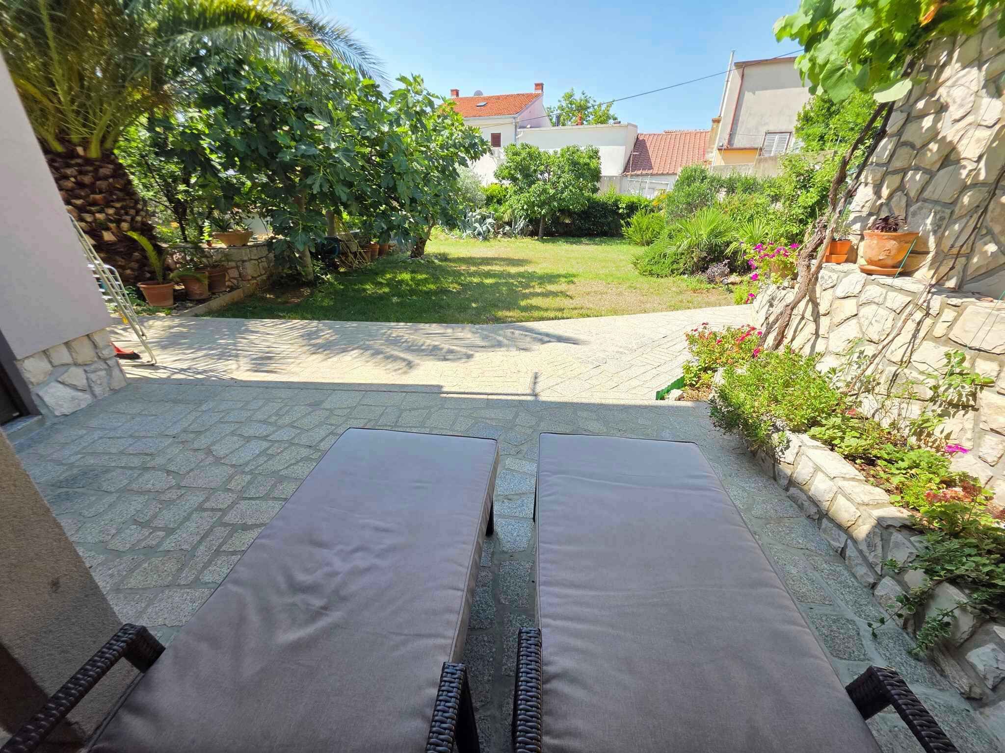 Een terras met twee loungestoelen, een stenen pad en een tuin met verschillende planten en een kleine boom.
