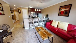 A living room with a red sofa, a patterned coffee table, and a kitchen area with a microwave and bar stools. - Maravilla Seawall Condo by AB Sea (Galveston)