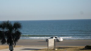 A coastal road with a white car parked on the side, a palm tree, and the ocean in the background. - Maravilla Seawall Condo by AB Sea (Galveston)