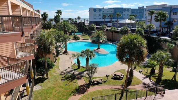 A pool area with palm trees, a picnic table, and a view of hotel buildings. - Maravilla Seawall Condo by AB Sea (Galveston)
