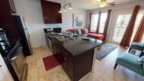 A compact kitchen with a central island, a dining area with a red sofa, and a view of the outdoors through large windows. - Maravilla Seawall Condo by AB Sea (Galveston)