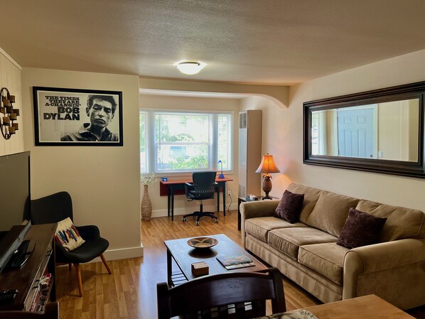A living room with a sofa, a coffee table, a desk with a chair, a lamp, and a poster on the wall. - Great downtown Sandpoint location! (Sandpoint)