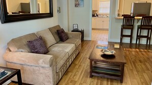 Ein Wohnzimmer mit einem beigen Sofa, einem Holztisch und einer durchgehenden Tür erkennbaren Küche.