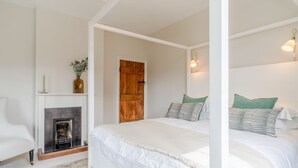 A bedroom with a fireplace, two beds with green pillows, a white chair, and a wooden door. - Court Cottage - sleeps 8 guests  in 4 bedrooms (Painswick)