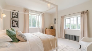 A bedroom with a large bed, two windows with curtains, a dresser, a chair, and a radiator.