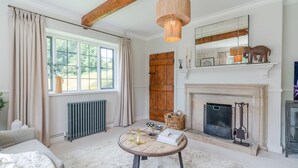 A room with a fireplace, a round wooden table with books and candles, a large window with curtains, and a wooden door. - Court Cottage - sleeps 8 guests  in 4 bedrooms (Painswick)