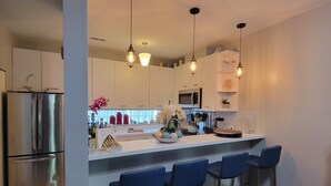 Eine moderne Küche mit einem Edelstahl-Kühlschrank, einer Mikrowelle und einem eingebauten Backofen. Drei Barhocker und eine weiße Arbeitsplatte mit verschiedenen Küchenutensilien.