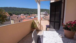 Un balcon avec une table et des chaises, donnant sur une ville avec des bâtiments aux toits rouges et une colline à l’horizon.