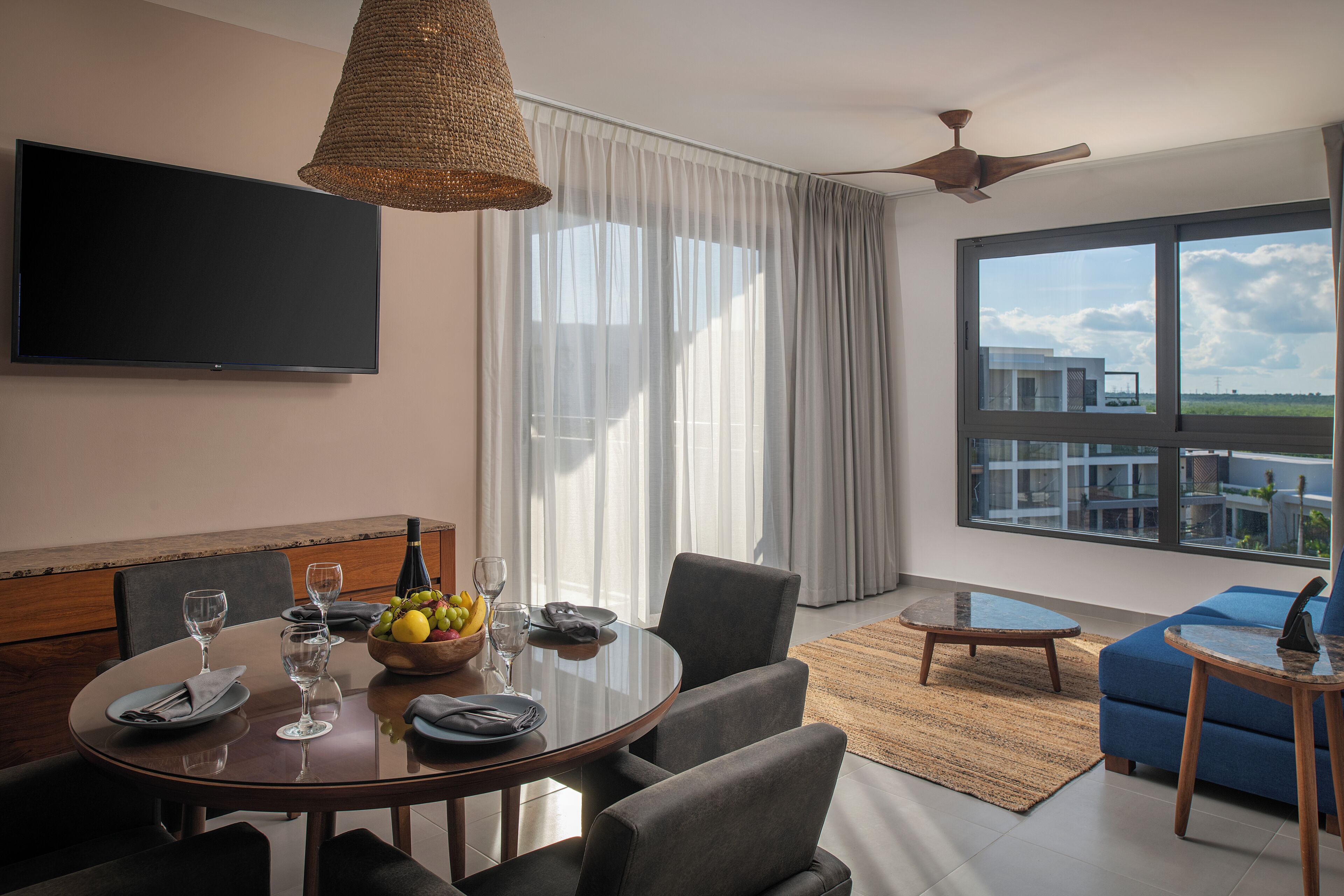 Una habitación de hotel moderna con zona de comedor, un televisor de pantalla plana en la pared, un ventanal con vistas y un ventilador de techo.