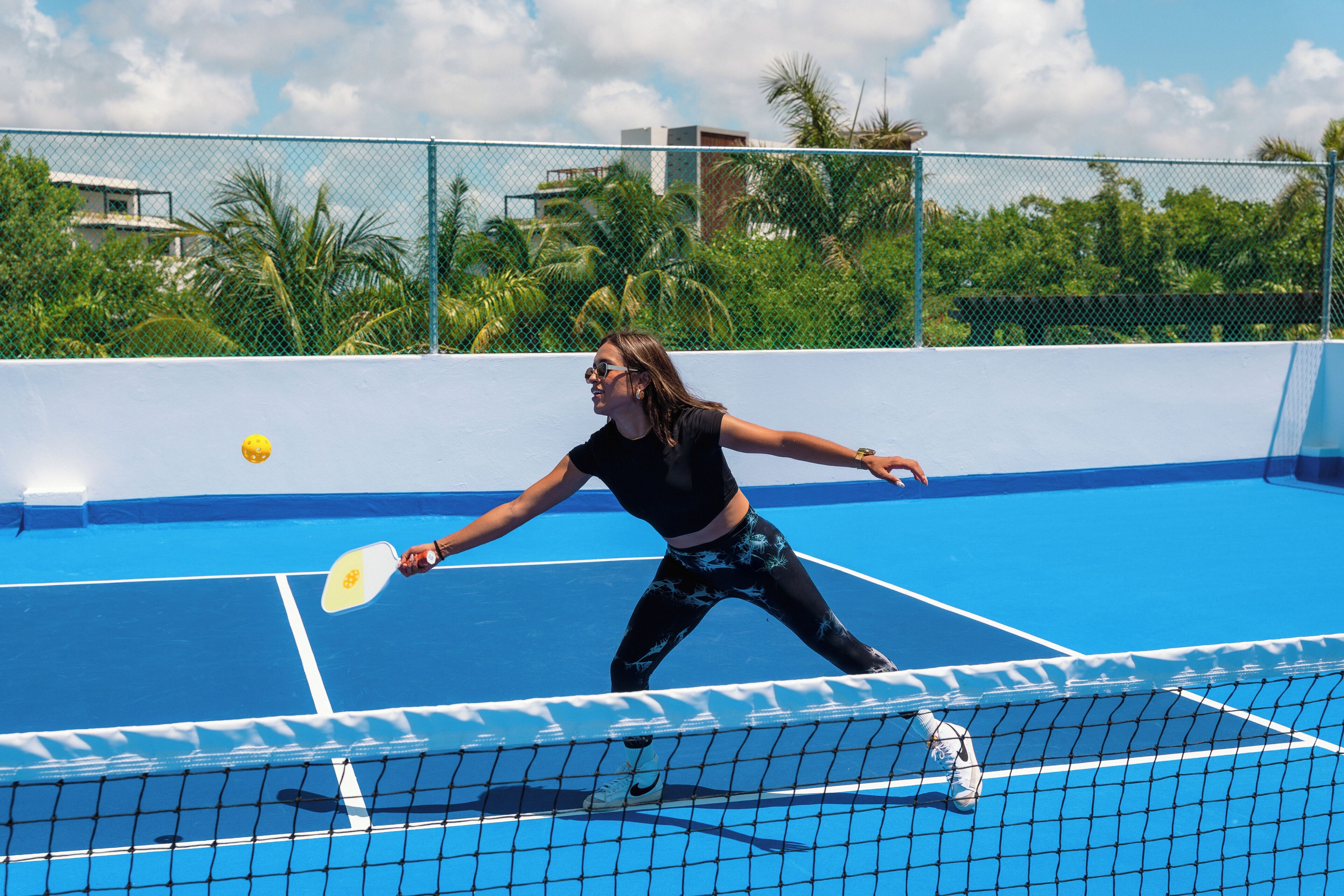 Una cancha de tenis con red, un jugador con camiseta negra y leggings estampados, y superficie de juego azul.