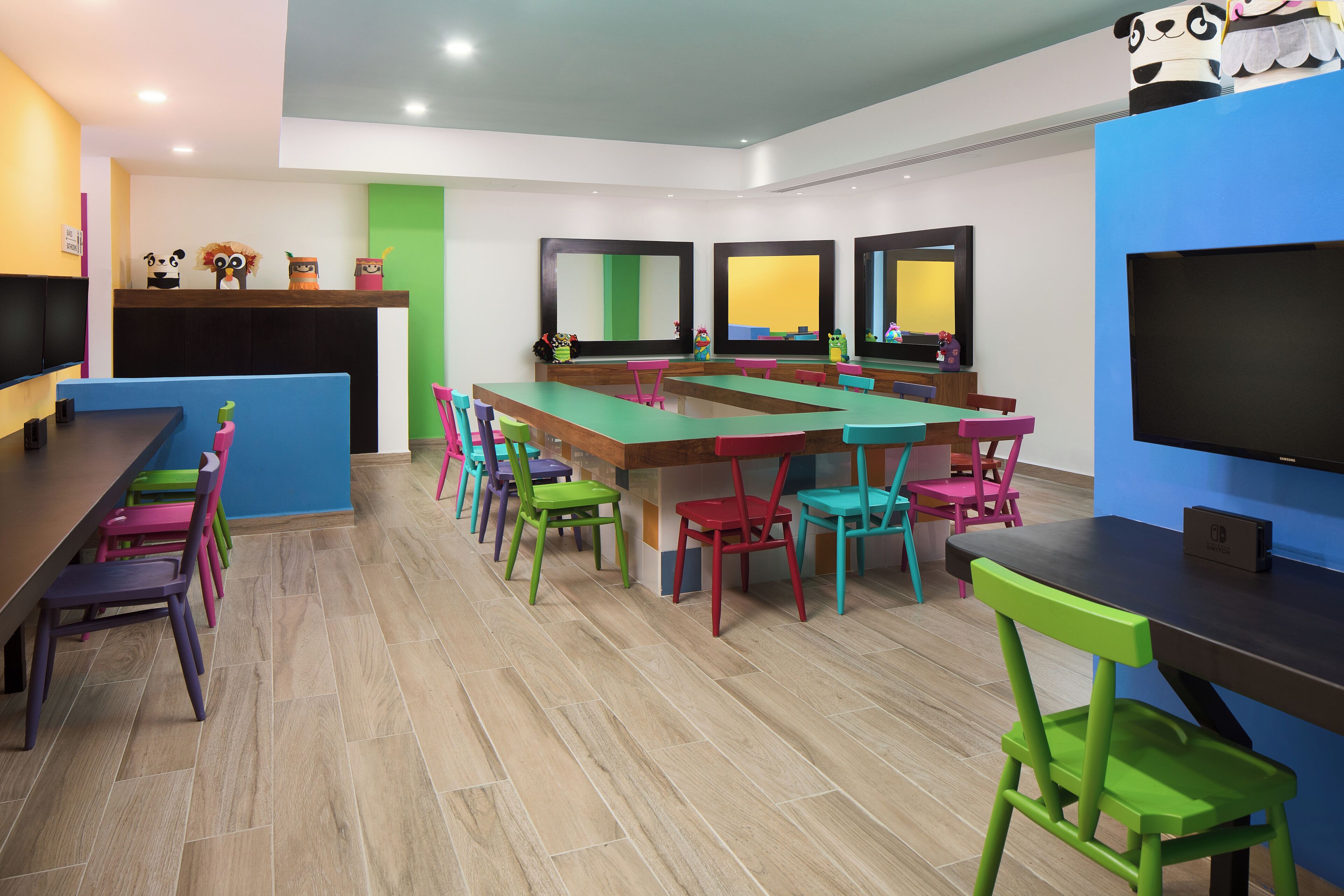 A children's play area with colorful chairs and tables, a large TV, and a panda-themed decoration.