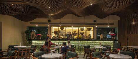 A restaurant with round tables and leather chairs, a bar area with a green wall and decorative items, and a wavy ceiling design.