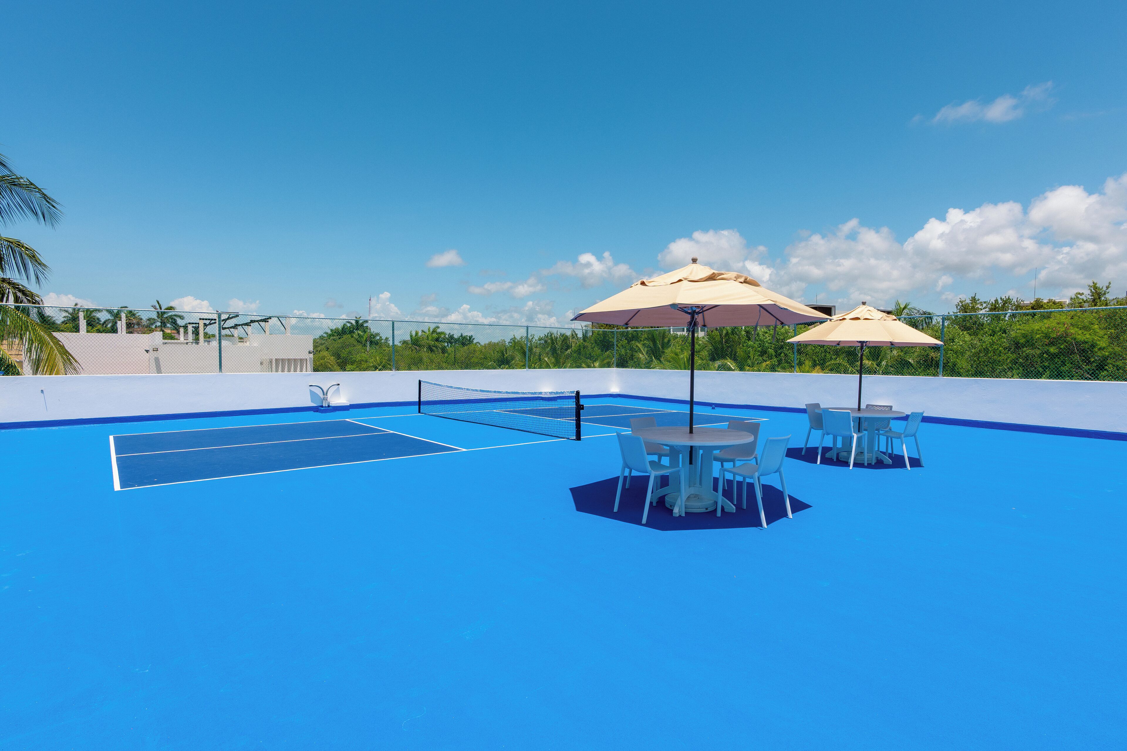 A blue tennis court with a net, surrounded by tables and chairs, and two umbrellas.
