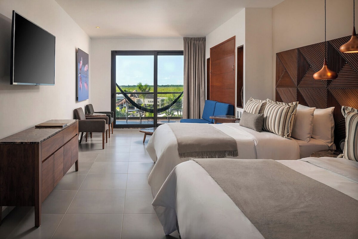 Une chambre d’hôtel équipée d’un lit, d’un bureau, d’une télévision et offrant une vue sur un hamac et la verdure environnante.