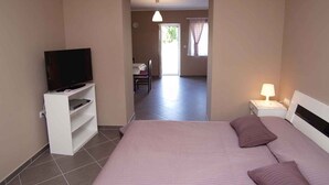 Una camera d'albergo con un letto, una televisione su un mobile, un comodino con una lampada e vista su una sala da pranzo attraverso una porta aperta.