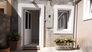 Un viuzza stretta con una porta bianca che dà su una stanza, una finestra con tenda e piante in vaso.