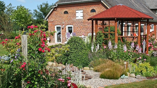 A brick house with a red roof, a wooden gazebo, and a garden with various flowers and plants.