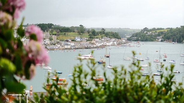 Une ville côtière où de nombreux bateaux sont amarrés le long du port, entourée d’une végétation luxuriante et de bâtiments historiques.