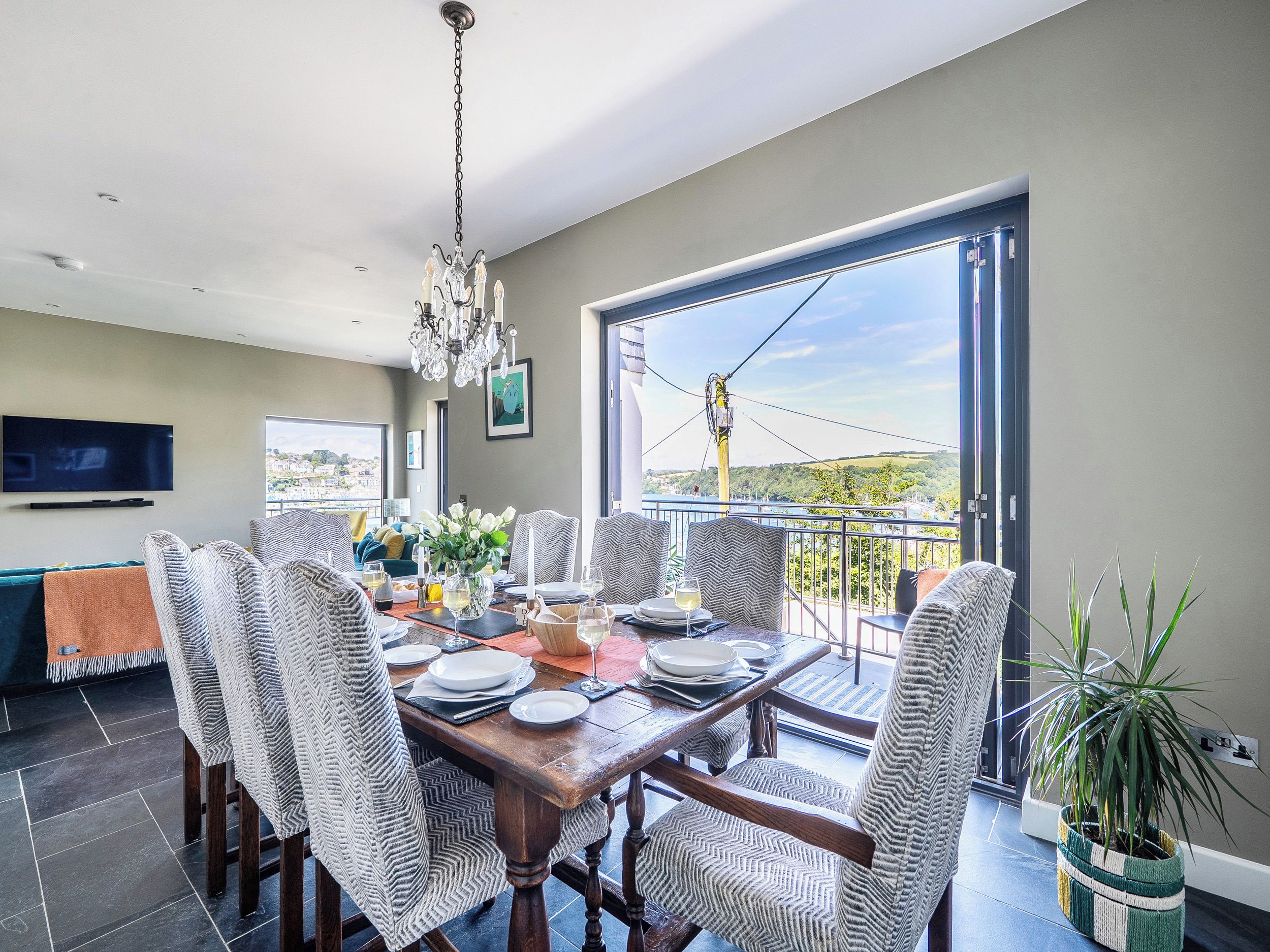 A dining area with a table set for four, surrounded by chairs, a chandelier, and a view through a large window.