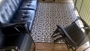 Een overdekt terras met een zwarte leren bank, een kamerkleed met dessin en een kleine ronde tafel met stoelen.