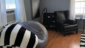 Een slaapkamer met een bed, een zwarte lederen fauteuil, een grijze poef, een zwart-wit gestreept kussen en een raam met gordijnen.