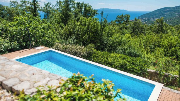 A rectangular swimming pool with clear blue water, surrounded by lush greenery and a view of the sea and distant hills. - Holiday home with pool, sauna and gym (Opatija)