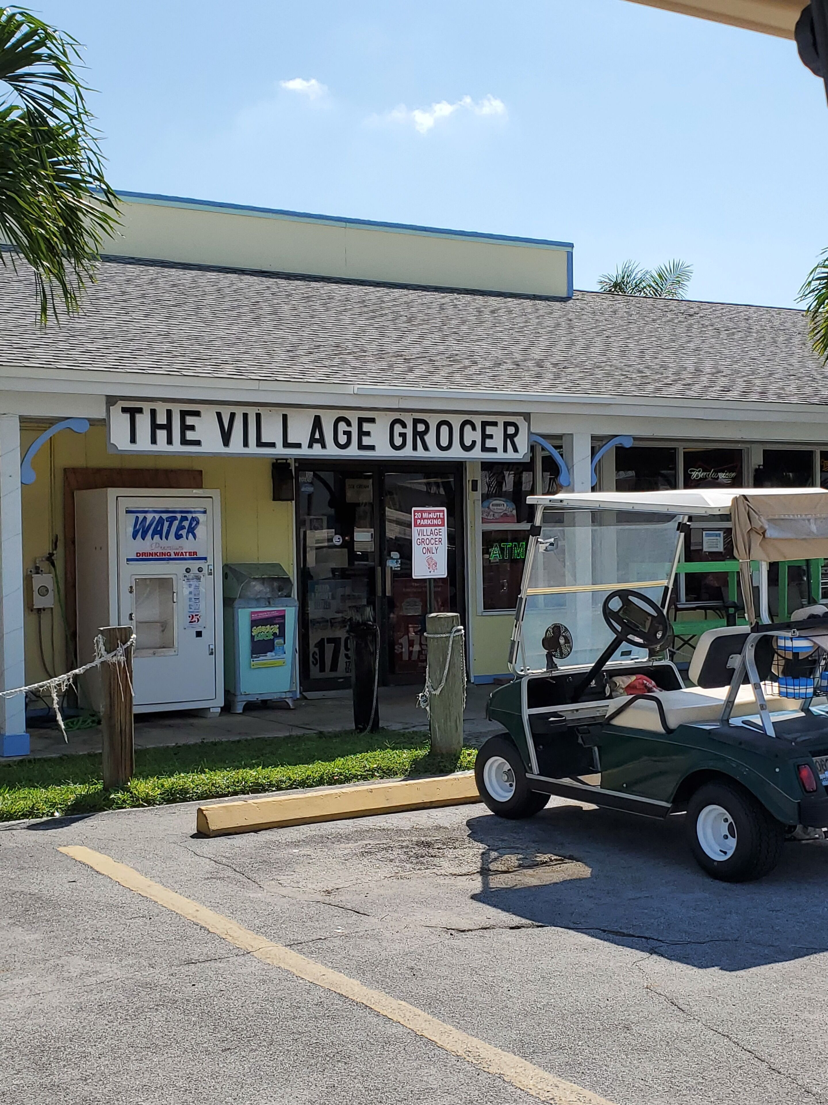 A village grocery store with a water dispenser, a 20-minute parking sign, and a golf cart parked in front.