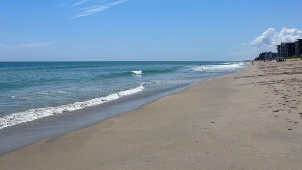 Ein Strand mit blauem Himmel, ruhiger See und Gebäuden im Hintergrund.
