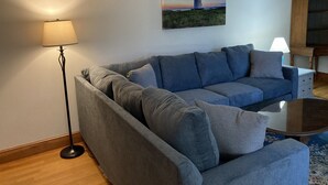 A living room with a grey sofa, a floor lamp, a coffee table, and a painting of a lighthouse on the wall. -  Downtown Edgartown Historic District. Walk to town and harbor. (Edgartown)