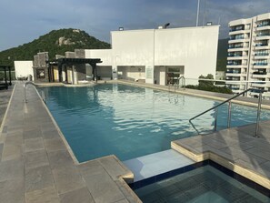 A swimming pool with a stone-tiled deck, a covered area with a grill, and a view of a mountain and residential buildings in the background. - Apartamento (Santa Marta)